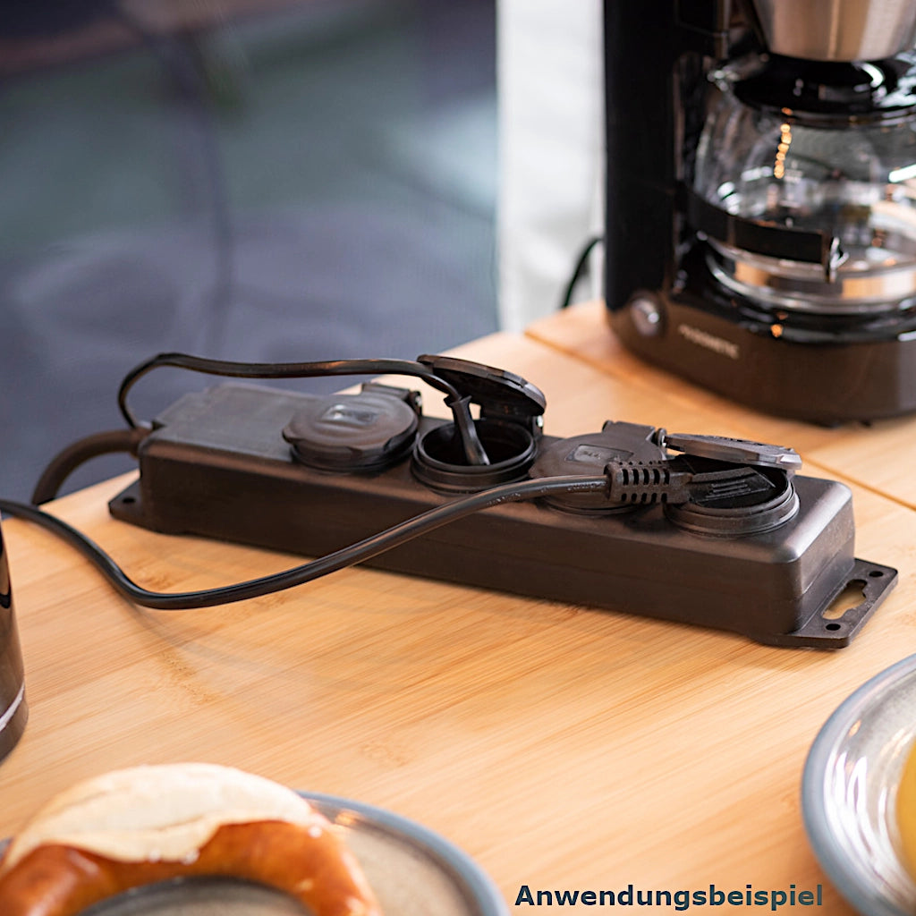 Eine schwarze Brennenstuhl Steckdosenleiste mit 2 eingesteckten Steckern liegt auf einem braunen Campingtisch