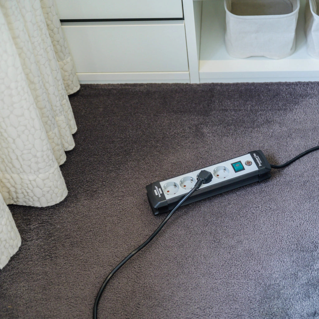 Eine Brennenstuhl Steckdosenleiste 4-fach mit Schalter liegt auf einem braunem Teppich vor einem Schrank