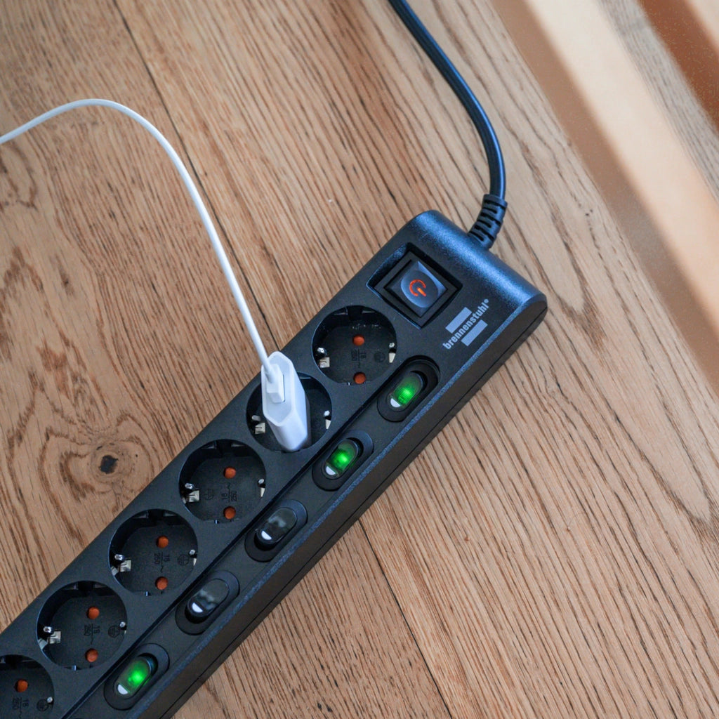Eine schwarze Brennenstuhl 6-fach Steckdosenleiste liegt auf einem Holzfußboden mit einem eingestecktem Stecker