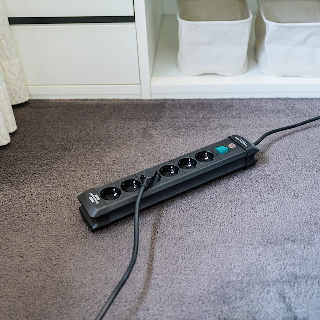 Eine schwarze Brennenstuhl Steckdosenleiste 6-fach mit Schalter liegt auf einem grauen Teppich vor einem Schrank