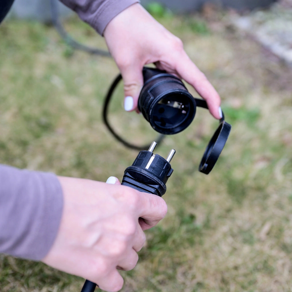 Ein Mann hockt auf einer grünen Wiese und hält eine schwarze Kupplung und einen schwarzen Stecker in je einer Hand