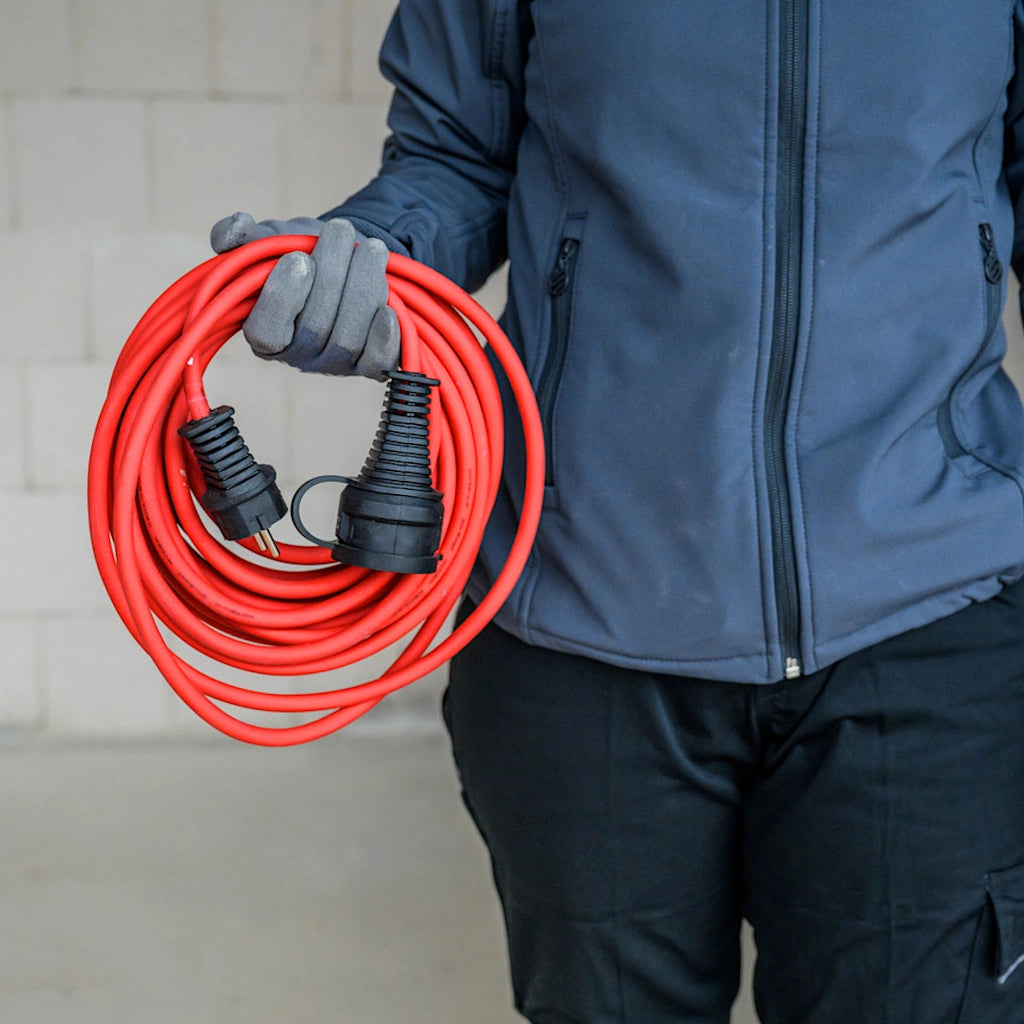 Ein Mann in blauer Arbeitskleidung hat ein aufgerolltes rotes Verlängerungskabel in seiner rechten Hand.