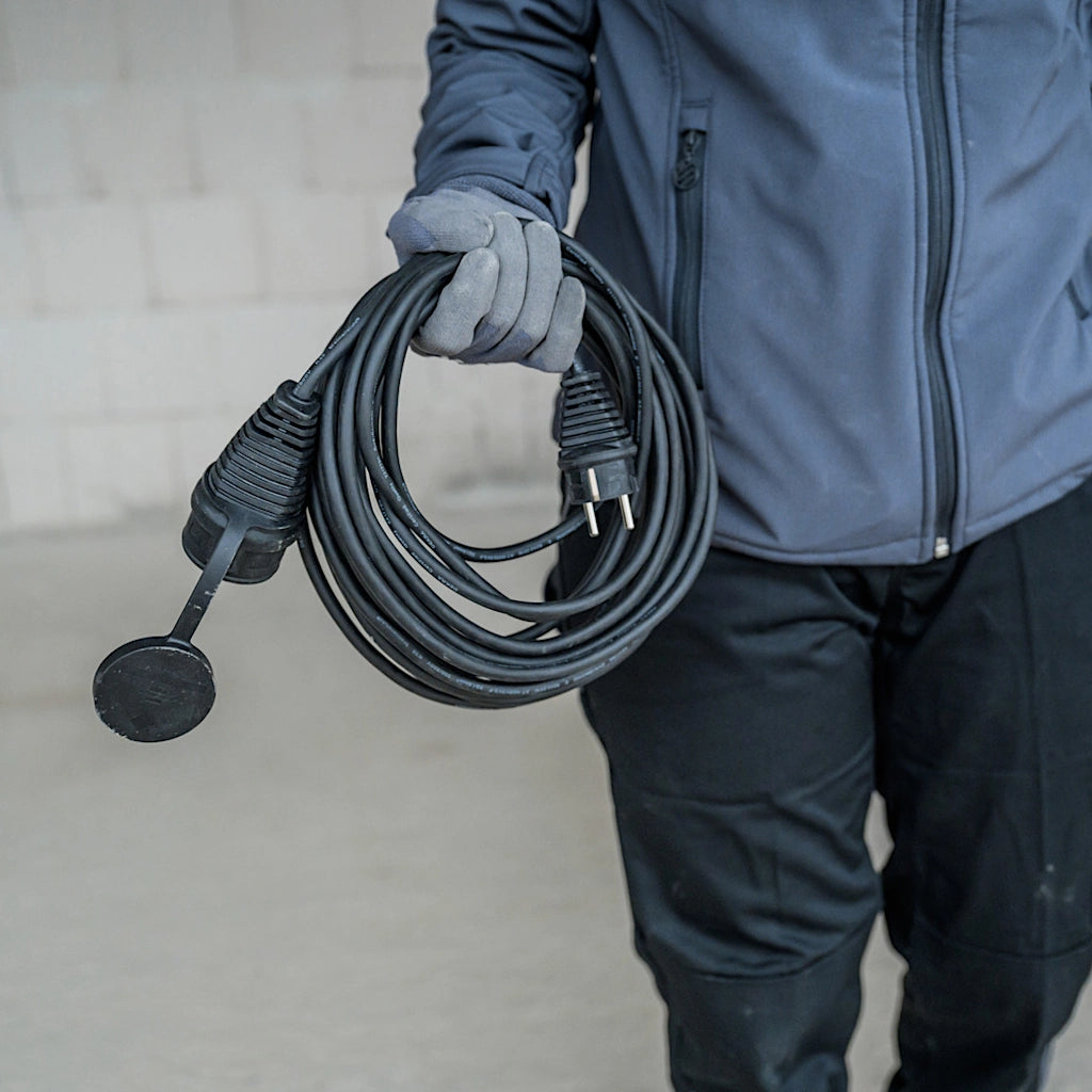 Ein Mann in blauer Arbeitskleidung hat ein aufgerolltes schwarzes Verlängerungskabel in seiner rechten Hand.