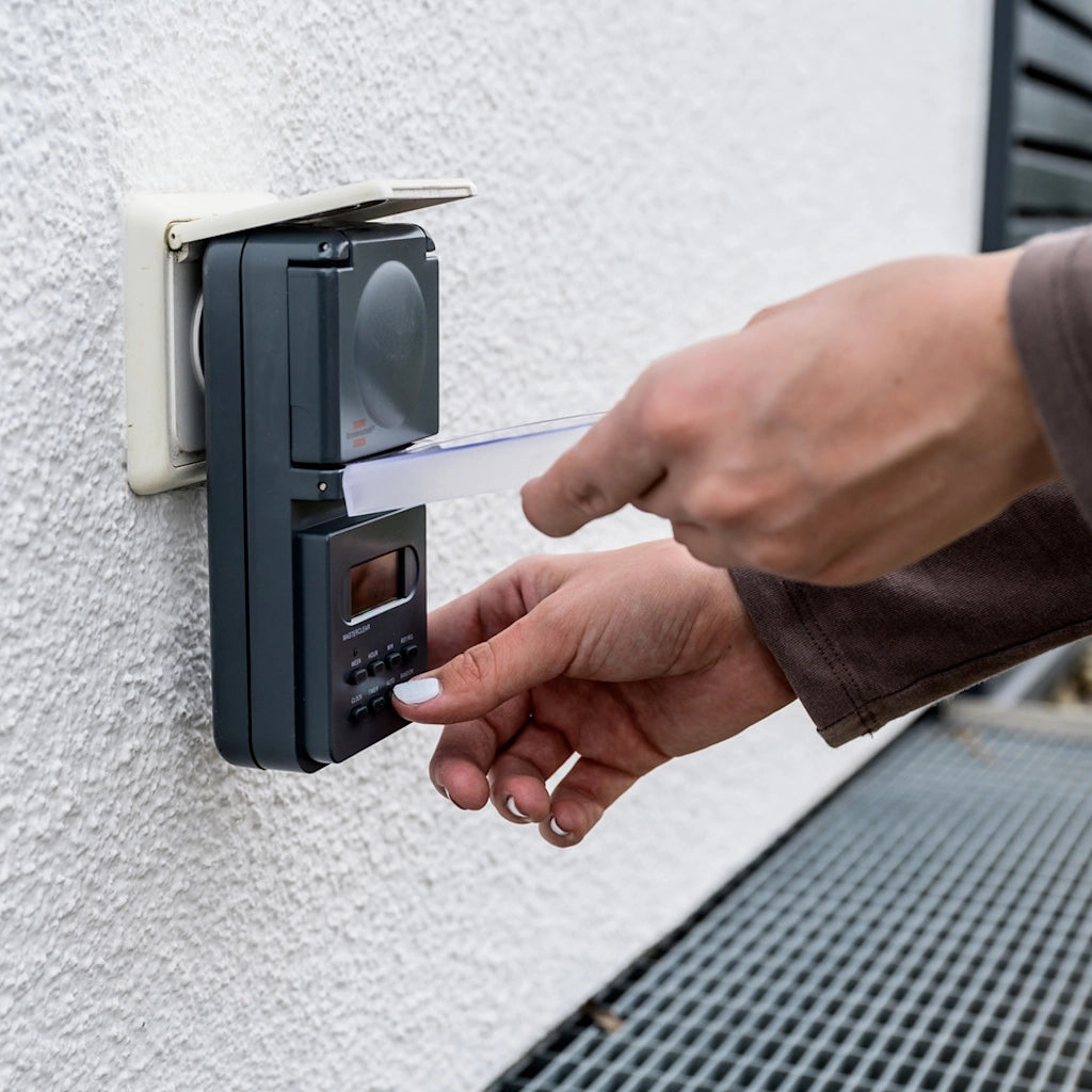 Eine Frau programmiert eine digitale Brennenstuhl Zeitschaltuhr in anthrazit die in einer Steckdose an einer Ausssenwand steckt