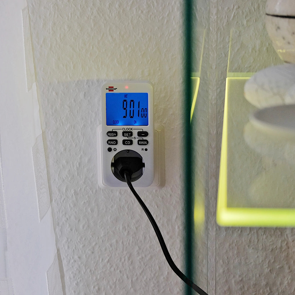 Eine weiße digitale Brennenstuhl Zeitschaltuhr mit beleuchteten Display steckt an einer weißen Wand in einer Steckdose