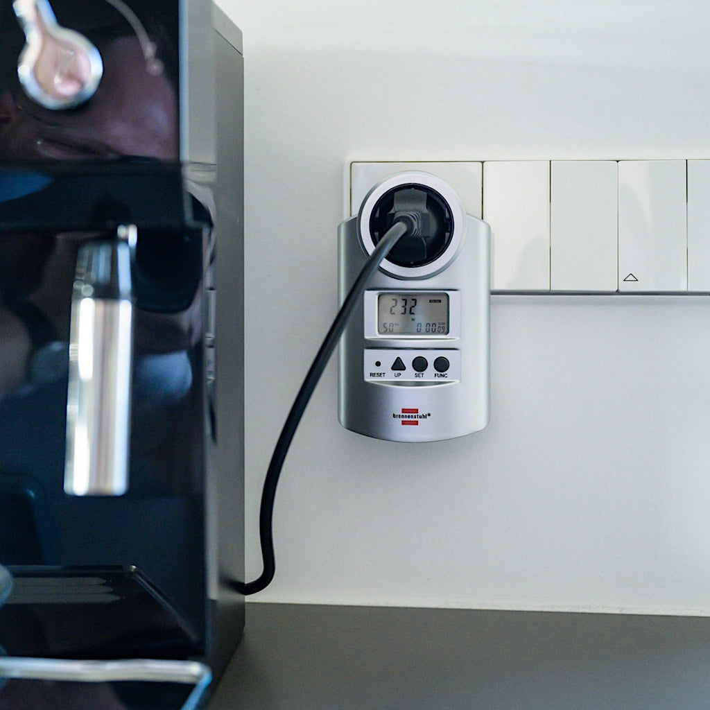 Ein silbernes Brennenstuhl Energiekostenmessgerät steckt an einer weißen Wand in einer Steckdose mit eingestecktem Stecker