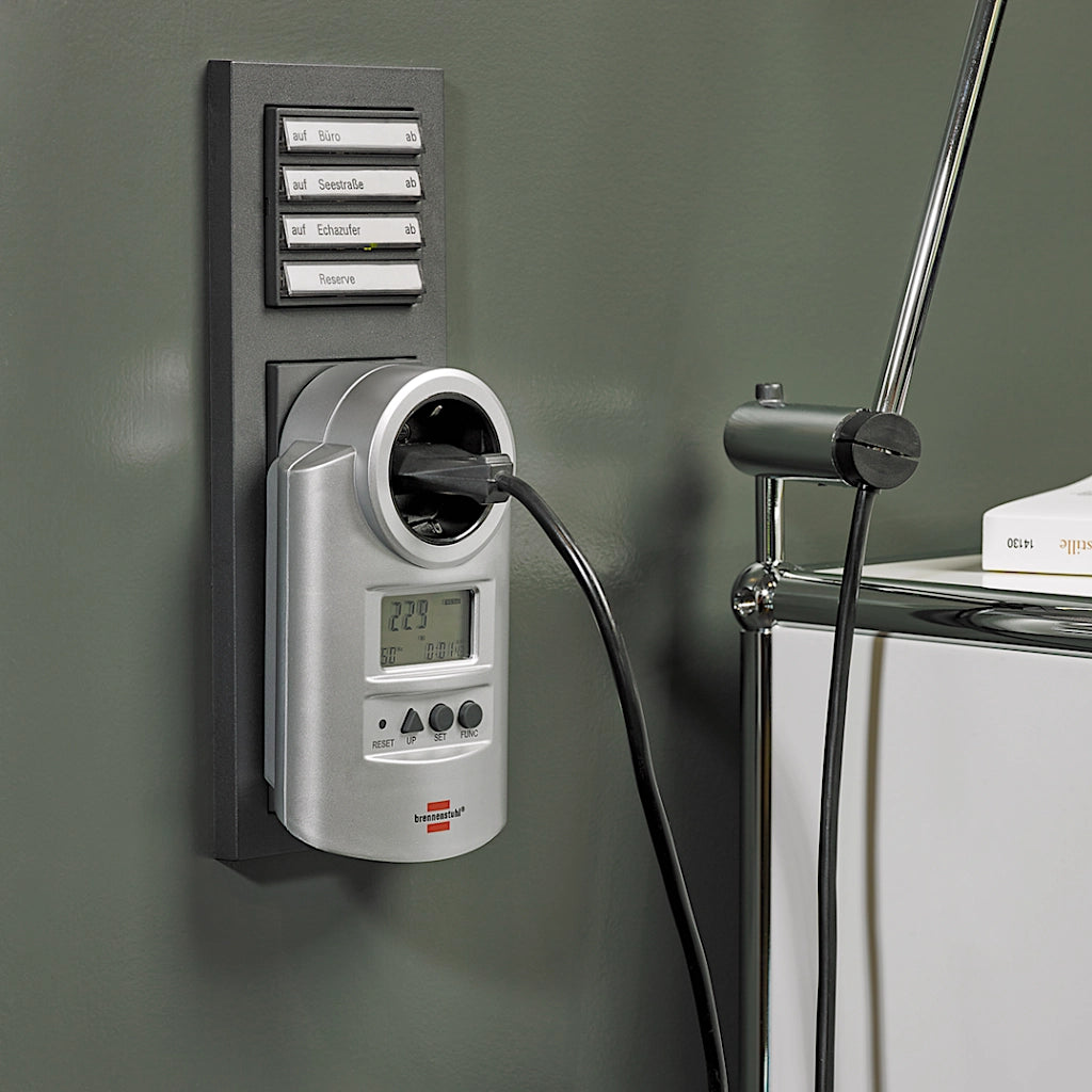 Ein silbernes Brennenstuhl Energiekostenmessgerät steckt an einer grünen Wand in einer Steckdose mit eingestecktem Stecker