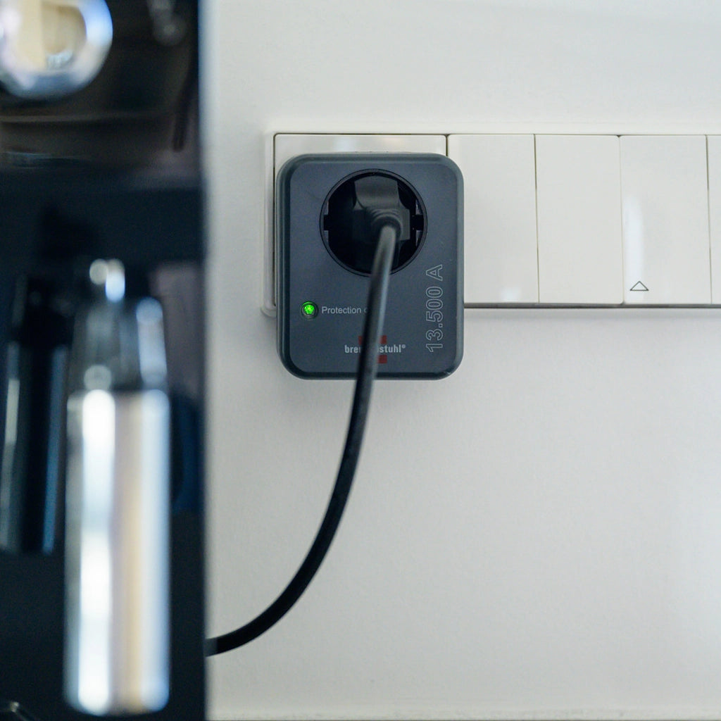 Ein Brennenstuhl Steckdosenadapter mit Überspannungsschutz steckt in einer Wandsteckdose mit eingestecktem Stecker einer Kaffeemachine.