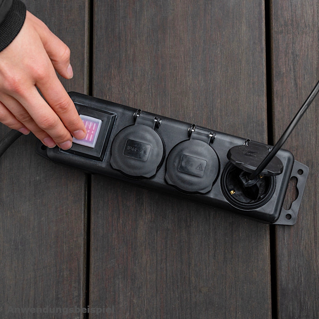 Eine Brennenstuhl Steckdosenleiste in schwarz liegt auf einem braunen Holzfußboden und eine Hand betätigt den Schalter