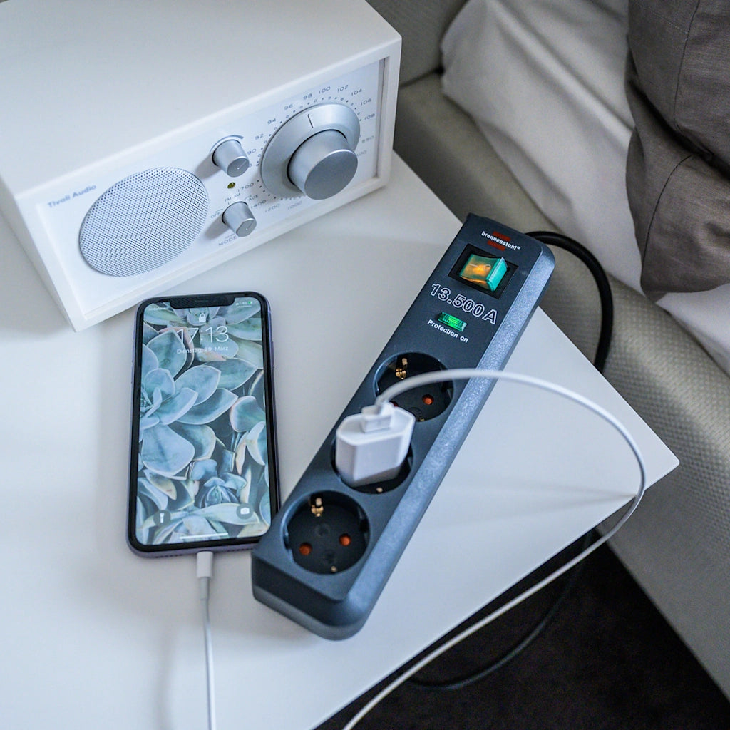 Eine Brennenstuhl 3-fach Steckdosenleiste in anthrazit mit Überspannungsschutz liegt neben einem Bett auf einem Tisch