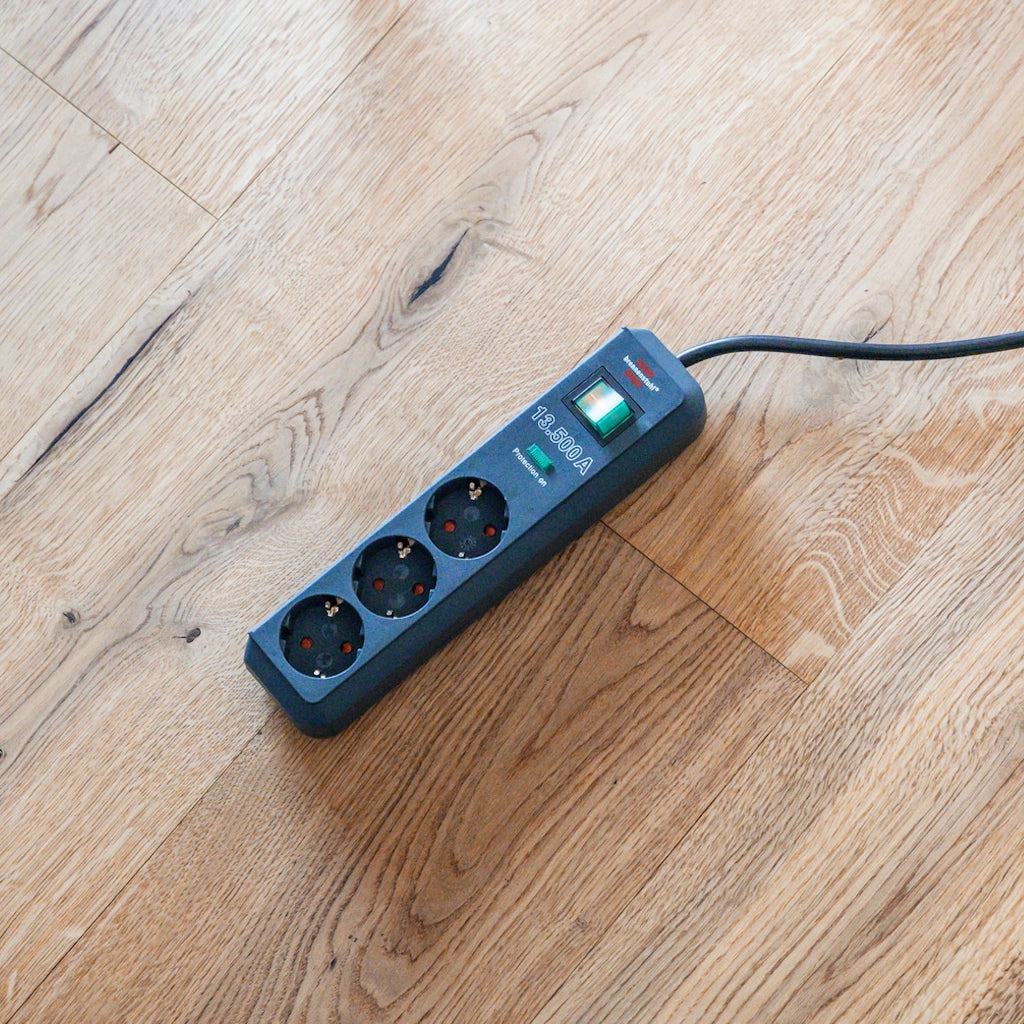 Eine Brennenstuhl 3-fach Steckdosenleiste in anthrazit mit Überspannungsschutz liegt auf einem Holzfußboden