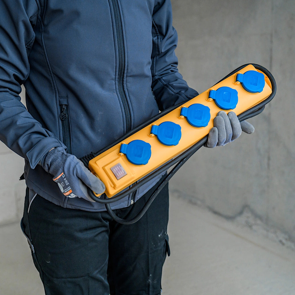 Ein Mann mit blauer Arbeitskleidung hält vor sich eine gelbe Steckdosenleiste mit blauen Deckeln