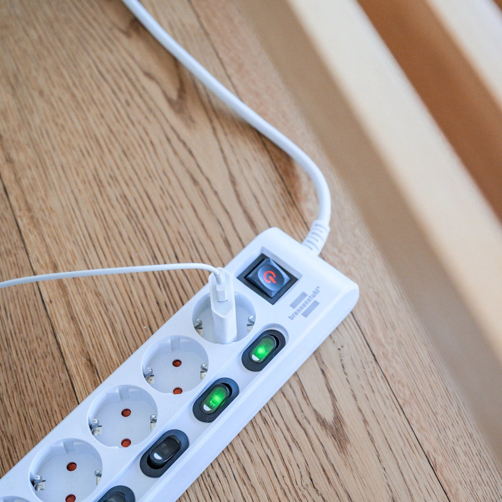 Eine weiße Brennenstuhl 6-fach Steckdosenleiste liegt auf einem Holzfußboden mit einem eingestecktem Stecker