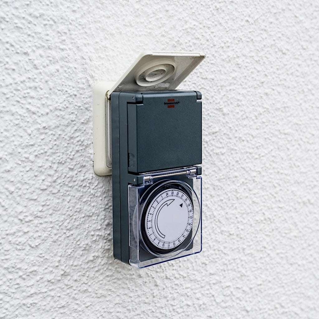 Eine mechanische Zeitschaltuhr in anthrazit von Brennenstuhl steckt in einer Unterputz Steckdose an einer weißen Wand im Aussenbereich