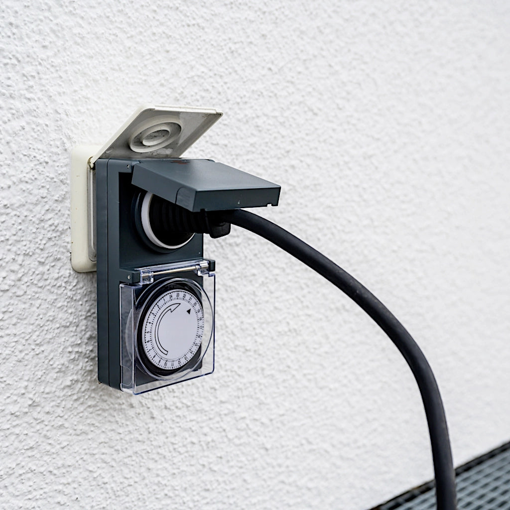 Eine mechanische Zeitschaltuhr in anthrazit von Brennenstuhl steckt in einer Unterputz Steckdose an einer weißen Wand im Aussenbereich mit einem angeschlossenem Stecker