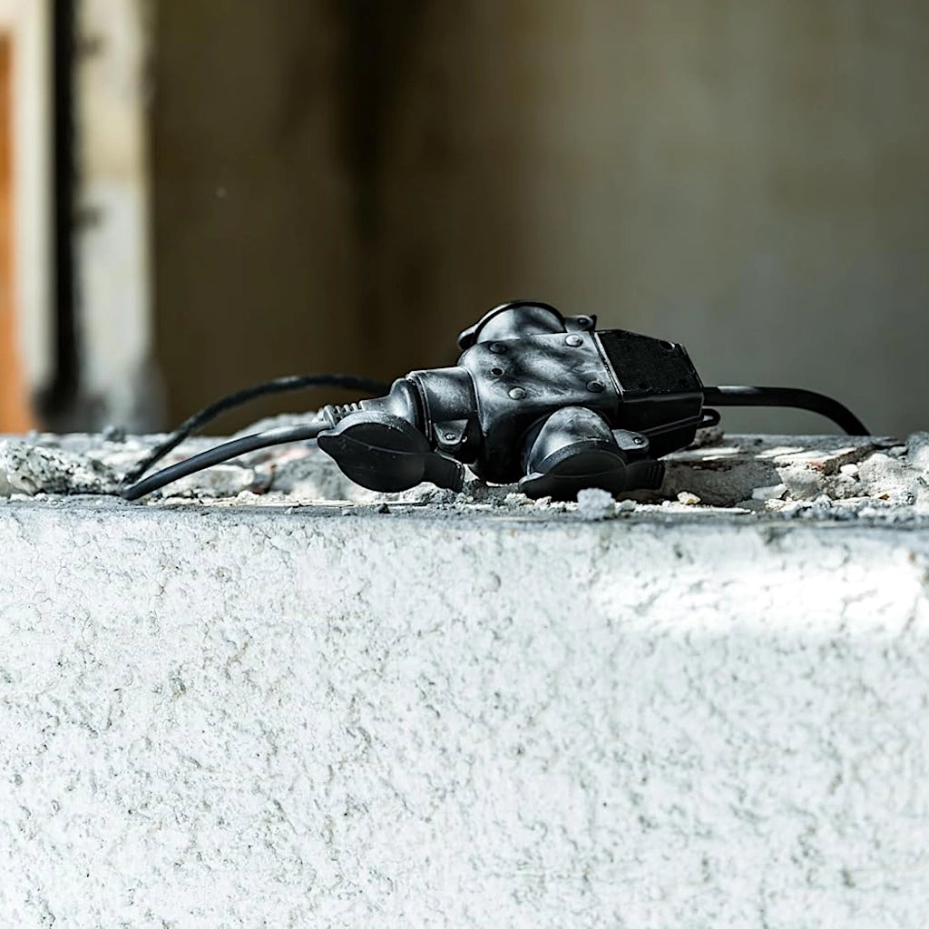 Eine 3-fach Hängekupplung aus Gummi in schwarz an der ein Stecker angeschlossen ist, liegt auf einer unfertigen Betonmauer.